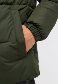Ciemnozielona kurtka puchowa z kieszenią na zamek z boku. Materiał jest gładki o matowym wykończeniu, a rękawy zakończone są elastycznymi mankietami.