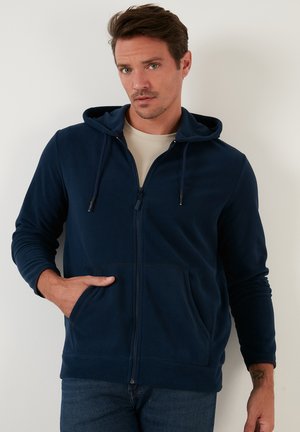 Blaue Kapuzenjacke mit Reißverschluss, Kapuze, Fronttaschen und strukturiertem Stoff. Verfügt über Zugbänder und gerippte Bündchen, kombiniert mit einem hellen T-Shirt.