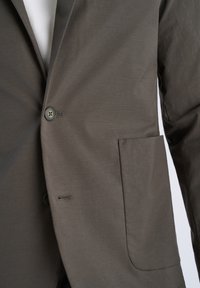 Καφέ μονόκουμπο σακάκι με δύο κουμπιά, μεγάλη μπροστινή τσέπη, φορεμένο πάνω από ένα λευκό πουκάμισο.