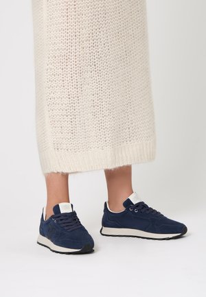 Sneaker in suede blu navy con suole e lacci in gomma bianca, dotati di un colletto imbottito e di un'etichetta con logo sulla linguetta. Indossati con una gonna in crema testurizzata.