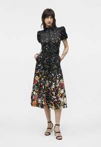 Femme portant une robe midi noire ceinturée avec des manches bouffantes et un imprimé floral et papillons multicolore, associée à des sandales noires à talons hauts.