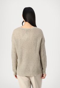 Personne aux longs cheveux foncés portant un pull en maille beige ample et un pantalon clair, vue de dos sur un fond uni.