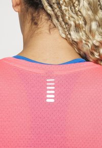 Różowa koszulka sportowa wykonana z teksturowanej tkaniny, z niebieskim wykończeniem przy dekolcie oraz charakterystycznymi białymi poziomymi paskami na plecach dla lepszej wentylacji.