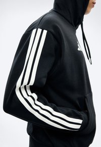 Άτομο που φοράει μαύρο φούτερ με λευκές τριπλές ρίγες στο μανίκι και τα χέρια στην μπροστινή τσέπη, εμφανιζόμενο από το πλάι.