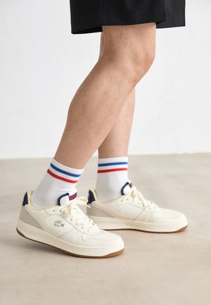 Sneakers bianche in pelle con pannelli laterali perforati, dettagli beige e suole in gomma marrone, abbinate a calzini bianchi con strisce rosse e blu.