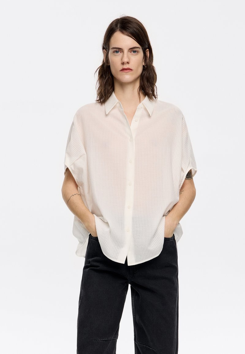 White textured button-up shirt with short sleeves and a loose fit, paired with dark jeans. Features a classic collar and front pockets.