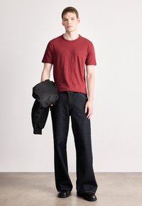 Κόκκινη κοντομάνικη μπλούζα με λογότυπο, συνδυασμένη με άνετο σκούρο τζιν. Το μοντέλο κρατάει ένα μαύρο bomber μπουφάν. Απλός, casual σχεδιασμός.