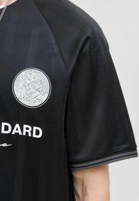 Maillot de sport noir à manches courtes avec des rayures verticales, texte blanc et un emblème rond détaillé présentant deux figures et des éléments floraux.