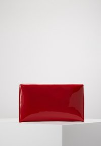 Pochette rouge brillante rectangulaire posée sur une surface angulaire blanche, avec un fond uni gris clair.