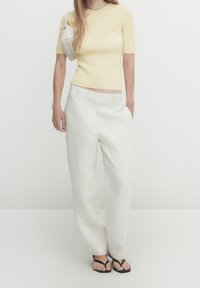 Light yellow ribbed short-sleeve top paired with loose-fitting white trousers, completed with black flat sandals and a small beige shoulder bag.