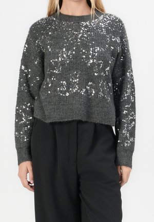 Femme portant un pull en maille gris foncé avec des sequins argentés et un pantalon noir uni taille haute, debout, les mains détendues.