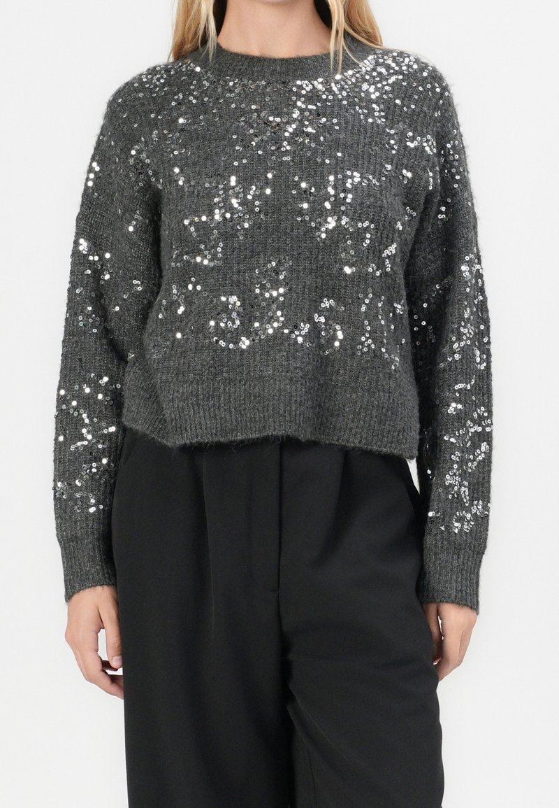 Femme portant un pull en maille gris foncé avec des sequins argentés et un pantalon noir uni taille haute, debout, les mains détendues.