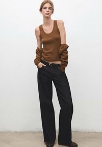 Top sin mangas marrón con canalé combinado con vaqueros negros de tiro alto y pierna ancha. La persona lleva un suéter marrón de gran tamaño caído sobre los hombros.