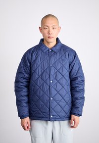Tamsiai mėlyna quilted striukė su apykakle, spaudėmis ir elastingomis rankovėmis. Ant krūtinės yra mažas logotipas, pagamintas iš blizgaus medžiagos.