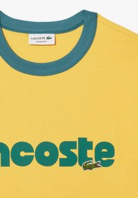 Żółta bawełniana koszulka z niebieskim ściągaczem, z wyraźnym zielonym nadrukiem "Lacoste" i małym haftowanym logo krokodyla.