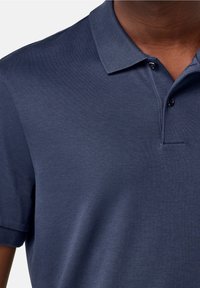 Námořnická modrá polo tričko vyrobené z hladkého materiálu; má klasický límec, dvouknoflíkovou légu a krátké rukávy.