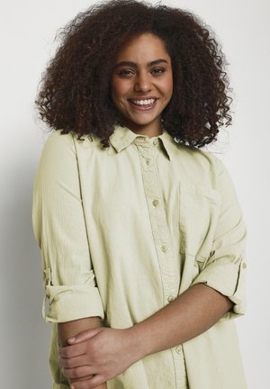 Tânără cu părul creț zâmbitoare, purtând o cămașă deschisă la culoare cu nasturi și mâneci suflecate, brațele ușor încrucișate în față.