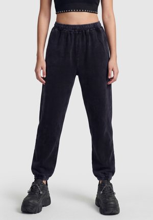 Osoba nosząca czarne spodnie dresowe z wysokim stanem, czarny crop top i czarne masywne sneakersy z detalem w postaci łańcucha z gwiazdką, stojąca na jednolitym tle.