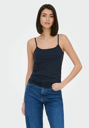 Woman wearing a black spaghetti strap tank top and blue jeans, standing with one hand in pocket against a plain light background.