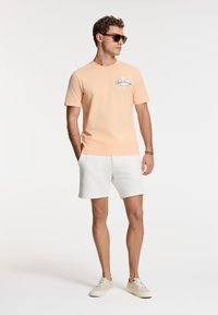 Lyse ferskenfarvet bomulds T-shirt med korte ærmer, med blå grafisk tekst. Kombineret med hvide shorts og lyse beige sneakers.