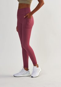 Leggings bordeaux avec une taille haute et une poche latérale, fabriqués dans un matériau extensible. Associés à des chaussures de sport blanches.