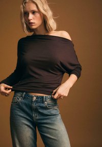 Top marrone off-shoulder realizzato in morbido tessuto, abbinato a jeans in denim chiaro a vita alta con chiusura a bottone e tasche.