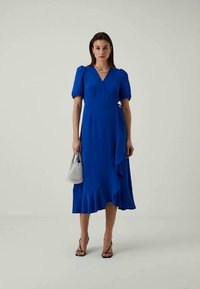 Nainen, jolla on päällään sininen kietaisumekko, jossa on puffi hihat ja röyhelöhelma, pitää kädessään pientä teksturoitua harmaata käsilaukkua, ja hänellä on jalassaan mustat remmikengät.