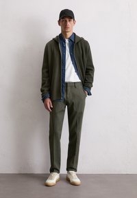 Dark green zip-up hoodie over a denim shirt, paired with olive trousers and cream sneakers. The cap is black with a logo.