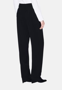 Zwarte wijde broek met een gladde textuur en hoge taille, gezien vanuit de achterkant. Gekleed met een witte blouse met lange mouwen en zwarte schoenen.