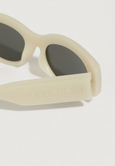 Beige napszemüveg sötét lencsékkel, kerek kerettel és lapos fülvéggel, mely oldalán "JIMMY CHOO" felirattal van gravírozva. Texturált felületi részlet.