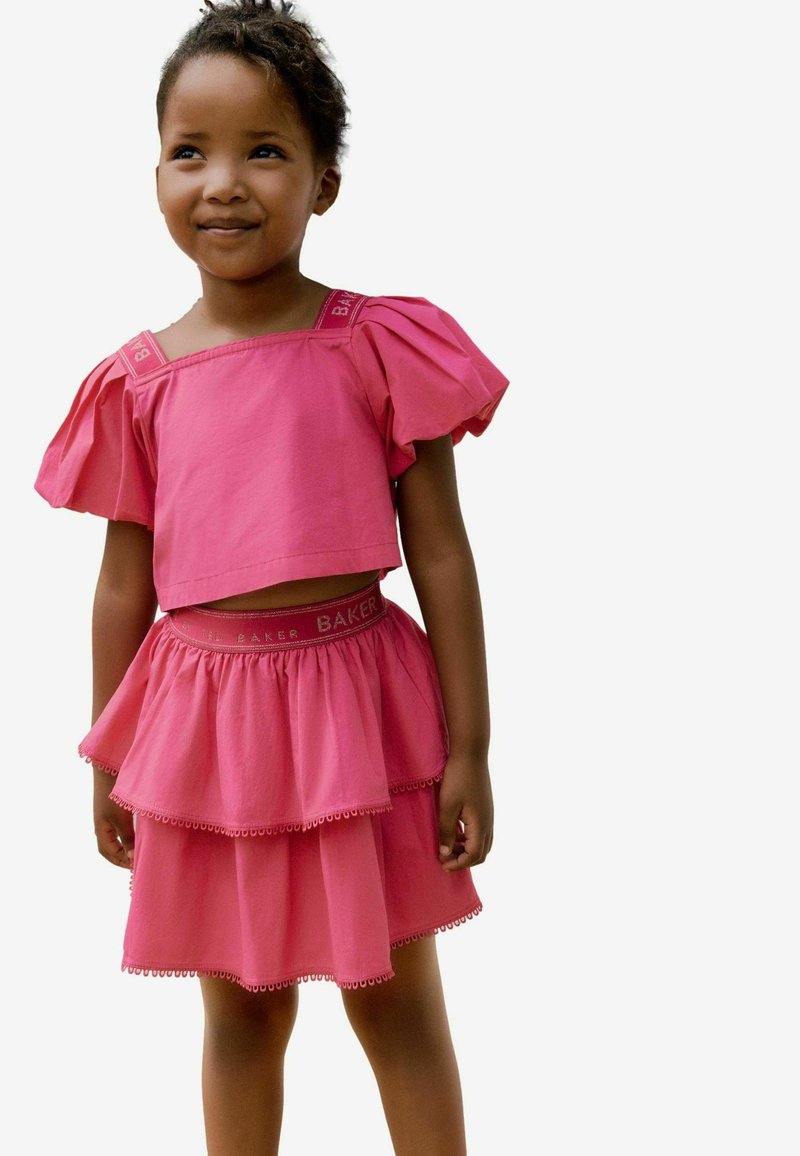 Lyserød cropped top med puffærmer og matchende tieret nederdel. Nederdelen har en dekorativ kant; toppen har fed skrift på stropperne.