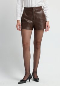 Donna che indossa pantaloni corti di pelle marrone, collant neri trasparenti, scarpe basse nere e una camicia bianca a maniche lunghe, in piedi contro uno sfondo semplice.