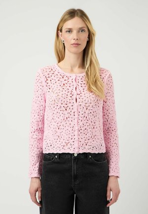 Donna bionda che indossa orecchini d'argento e un top a maniche lunghe in crochet floreale rosa chiaro abbinato a jeans neri a vita alta.