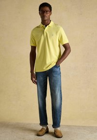 Homme portant un polo jaune, un jean bleu, des mocassins beige et des lunettes, debout avec une main dans la poche contre un fond beige uni.