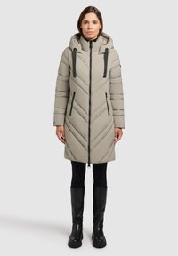 Giacca piumino beige a motivo chevron con cappuccio, chiusura con zipper e dettagli neri. Il modello indossa leggings neri e stivali neri alti fino al ginocchio.