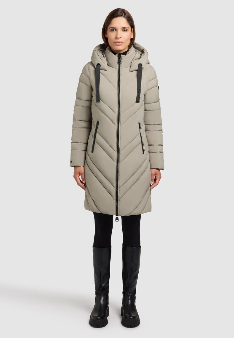 Giacca piumino beige a motivo chevron con cappuccio, chiusura con zipper e dettagli neri. Il modello indossa leggings neri e stivali neri alti fino al ginocchio.