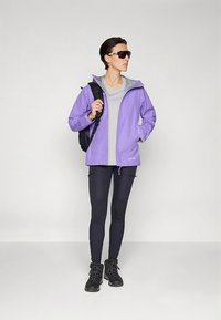 Jasnofioletowa wodoodporna kurtka z szarymi detalami, szara koszula, czarne legginsy i czarne buty turystyczne, w towarzystwie czarnego plecaka.