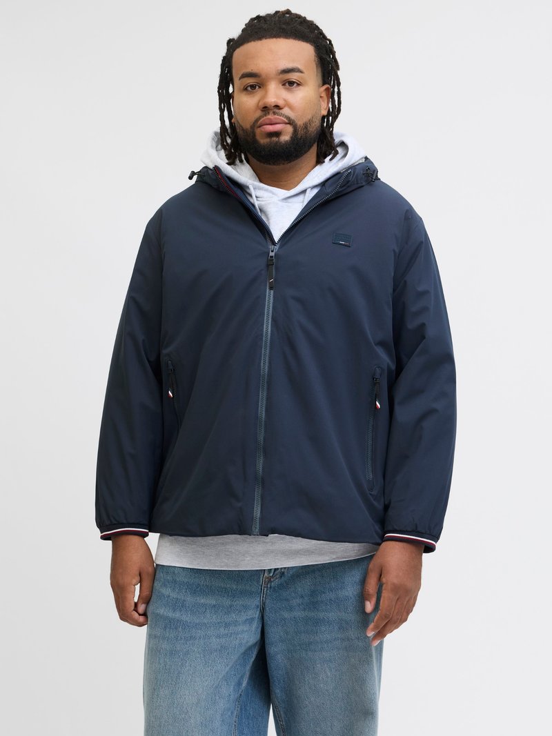 Giacca leggera blu navy con cappuccio, dotata di tasche con cerniera e dettagli a contrasto sui polsini. Indossata sopra un felpa grigia.