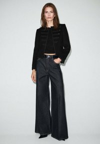 Czarna kurtka w stylu wojskowym z złotymi guzikami, zestawiona z wysokim stanem ciemnych dzwonowatych dżinsów. Gładki materiał, dopasowany krój, minimalistyczny design.