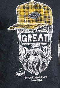 T-shirt noir présentant un graphique d'un visage barbu portant une casquette en tartan jaune. Un texte blanc en gras lit "SUPER" et "Trop Stylé !" en dessous.
