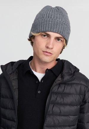 Szary dzianinowy czapka z prążkowanym teksturem. Model nosi czarną kurtkę puchową na czarnym polo z białą koszulą pod spód.