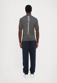 Grå t-skjorte med en vertikal hvit logo på baksiden, kombinert med marineblå joggebukser og hvite sko. Stoffet ser glatt og tett ut.