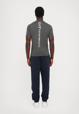 Gray t-shirt with a vertical white logo on the back, paired with navy sweatpants and white shoes. The fabric appears smooth and fitted.