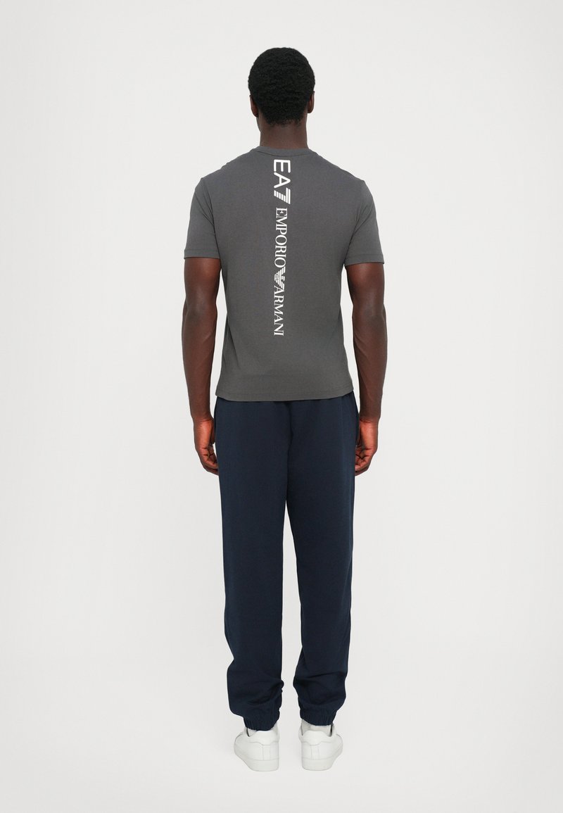 Grå t-skjorte med en vertikal hvit logo på baksiden, kombinert med marineblå joggebukser og hvite sko. Stoffet ser glatt og tett ut.
