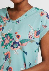 Blouse à manches courtes et décolleté en V de couleur turquoise clair avec des motifs floraux en rose, bleu et vert. Tissu doux avec une texture lisse.