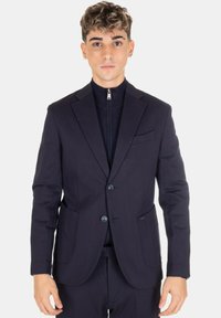Giovane uomo che indossa un blazer blu navy aderente con due bottoni sopra un top nero zip, in piedi contro uno sfondo bianco semplice.