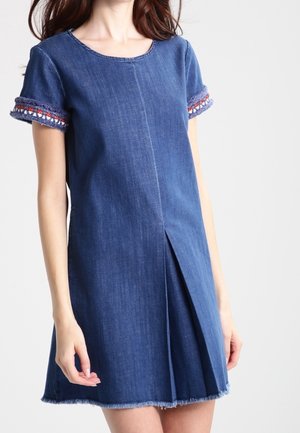 Kvinde iført en kortærmet denimkjole med broderet kant på ærmerne og en detaljeret forstykke med læg.