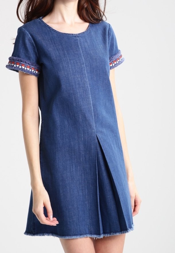 Femme portant une robe en jean à manches courtes avec un bord brodé sur les manches et un détail de pli sur le devant.
