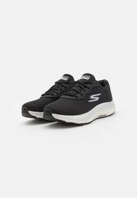 Schwarze Sportschuhe mit Obermaterial aus Netzgewebe, weißer Zwischensohle und schwarzem Gummiaußensohle, mit Logo an der Seite und Schnürdesign.