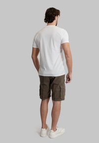 T-shirt bianca in cotone con maniche corte, abbinata a pantaloni cargo verde oliva e sneaker bianchi, vista da dietro su uno sfondo grigio.